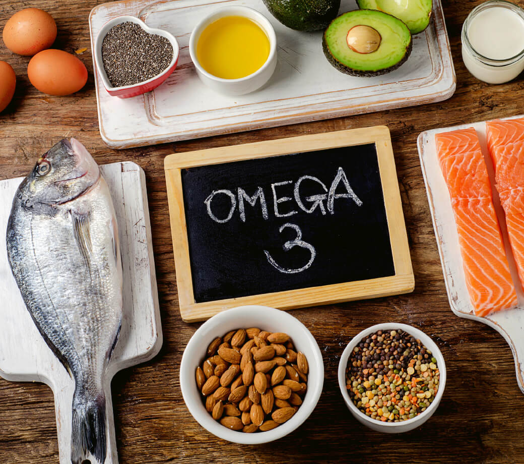 Natural Pharmaceuticals OmegaMarine™ Premium Az omega-3 zsírsavak különféle forrásai egy faasztalon - friss lazac, egész hal, tojás, avokádó, olívaolaj, chia mag, diófélék, tej, valamint hüvelyesek és gabonafélék keveréke. Középen egy tányér az „OMEGA 3” felirattal.